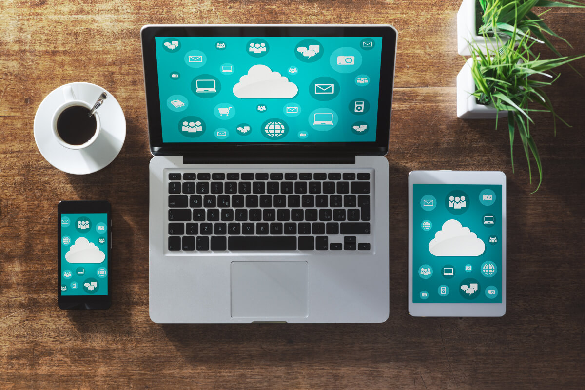 A mobile phone, laptop, and tablet lined up next to each other on a table next to a cup of coffee, each displaying a a 2D graphic of a cloud over a bluish-green background to represent Cloud technology.