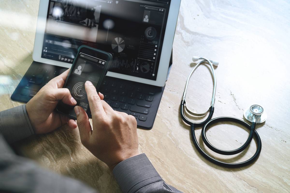 A doctor using a cellphone and laptop with a stethoscope resting on his desk